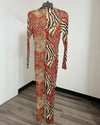 A long-sleeve, high-neck, full-length jumpsuit with an abstract animal-print pattern in rust red, beige, and black, hanging on a black hanger against a plain white wall.
