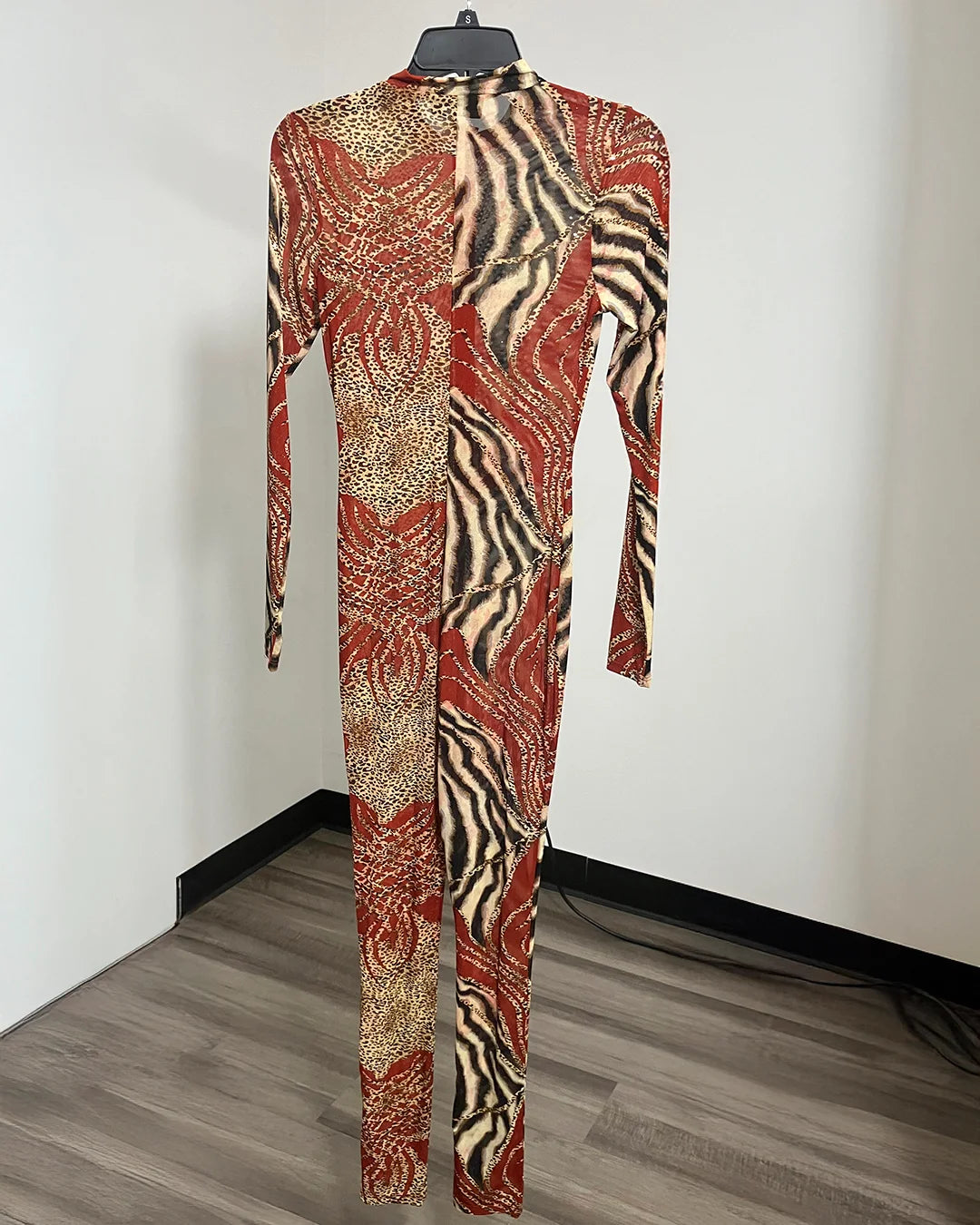 A long-sleeve, high-neck, full-length jumpsuit with an abstract animal-print pattern in rust red, beige, and black, hanging on a black hanger against a plain white wall.
