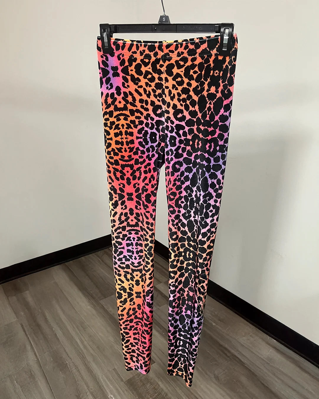 A pair of high-waisted leggings with a vibrant neon leopard print pattern, hanging on a black hanger against a plain wall.