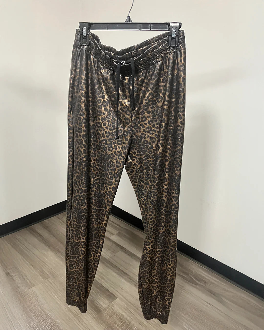 Leopard print jogger pants hanging on a black hanger against a plain off-white wall with a wood-look floor.