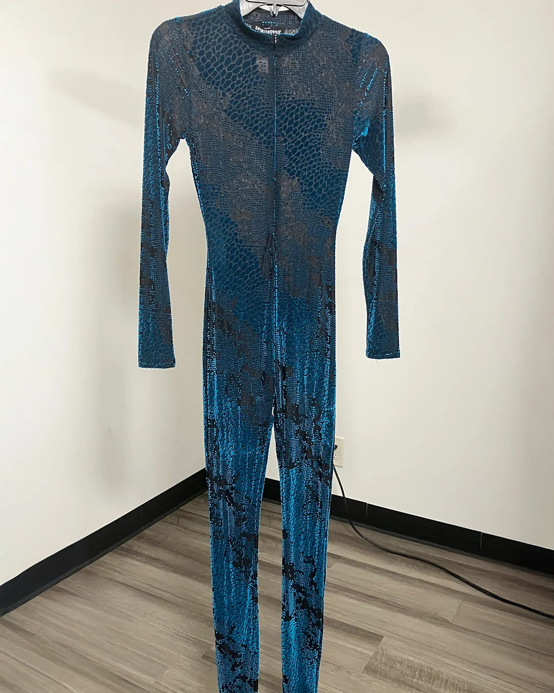 A long-sleeve, full-length jumpsuit with a blue and black snakeskin pattern, displayed on a hanger against a plain indoor wall.