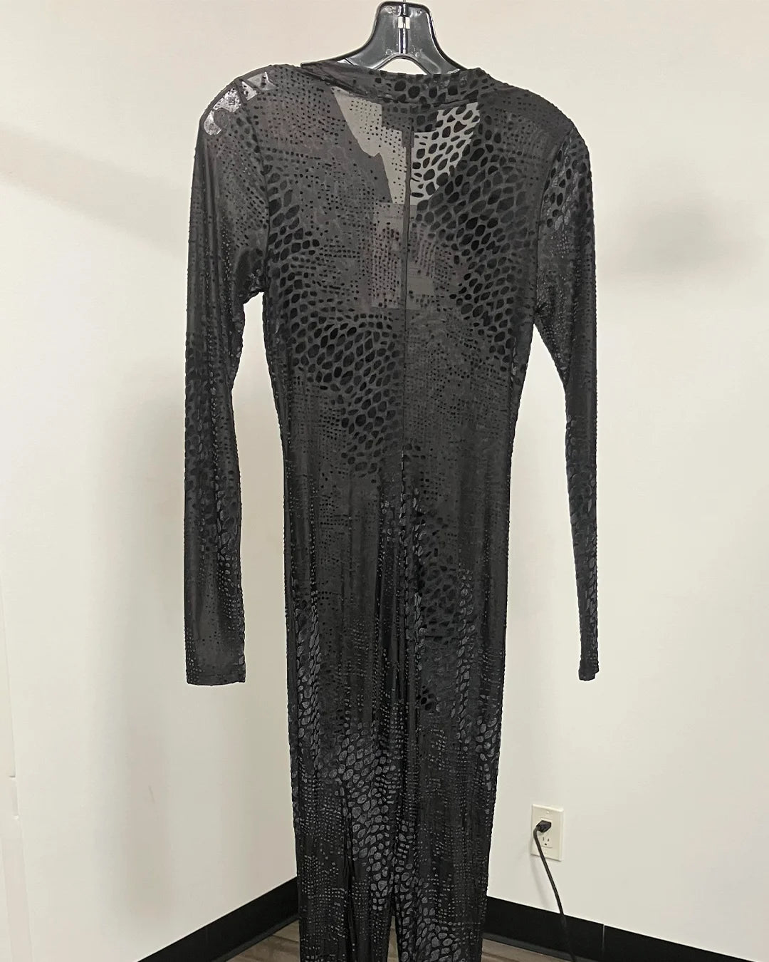 A long-sleeve black mesh jumpsuit with a textured, reptile-like pattern, hanging on a black hanger against a plain white wall.