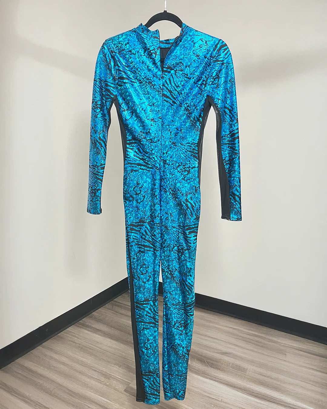 A long-sleeve one-piece jumpsuit in a vibrant blue and black abstract animal print pattern, hanging on a black hanger against a plain wall with wood flooring.