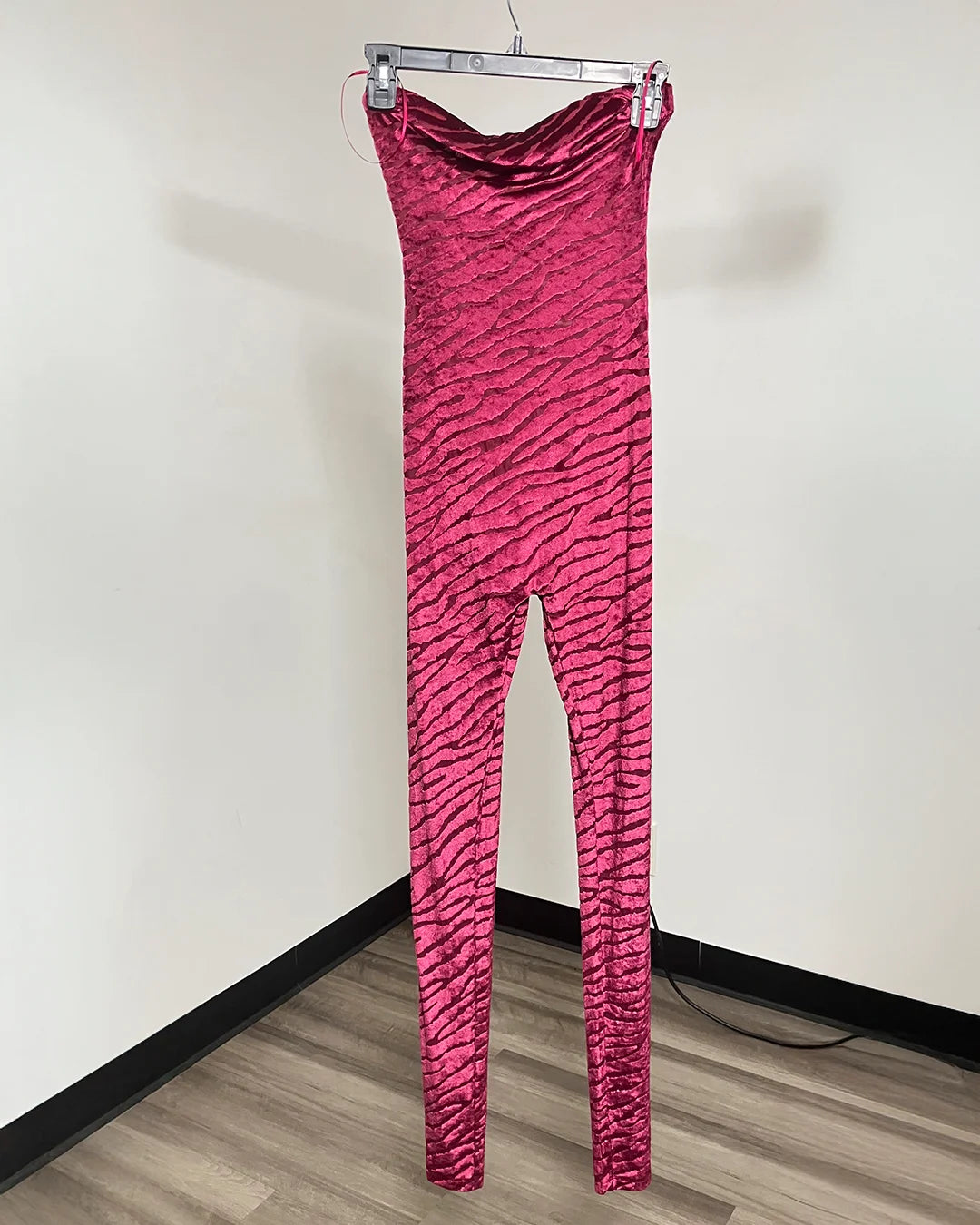 A bright pink zebra-print catsuit hanging from a metal hanger against a plain off-white wall and wood-look floor.