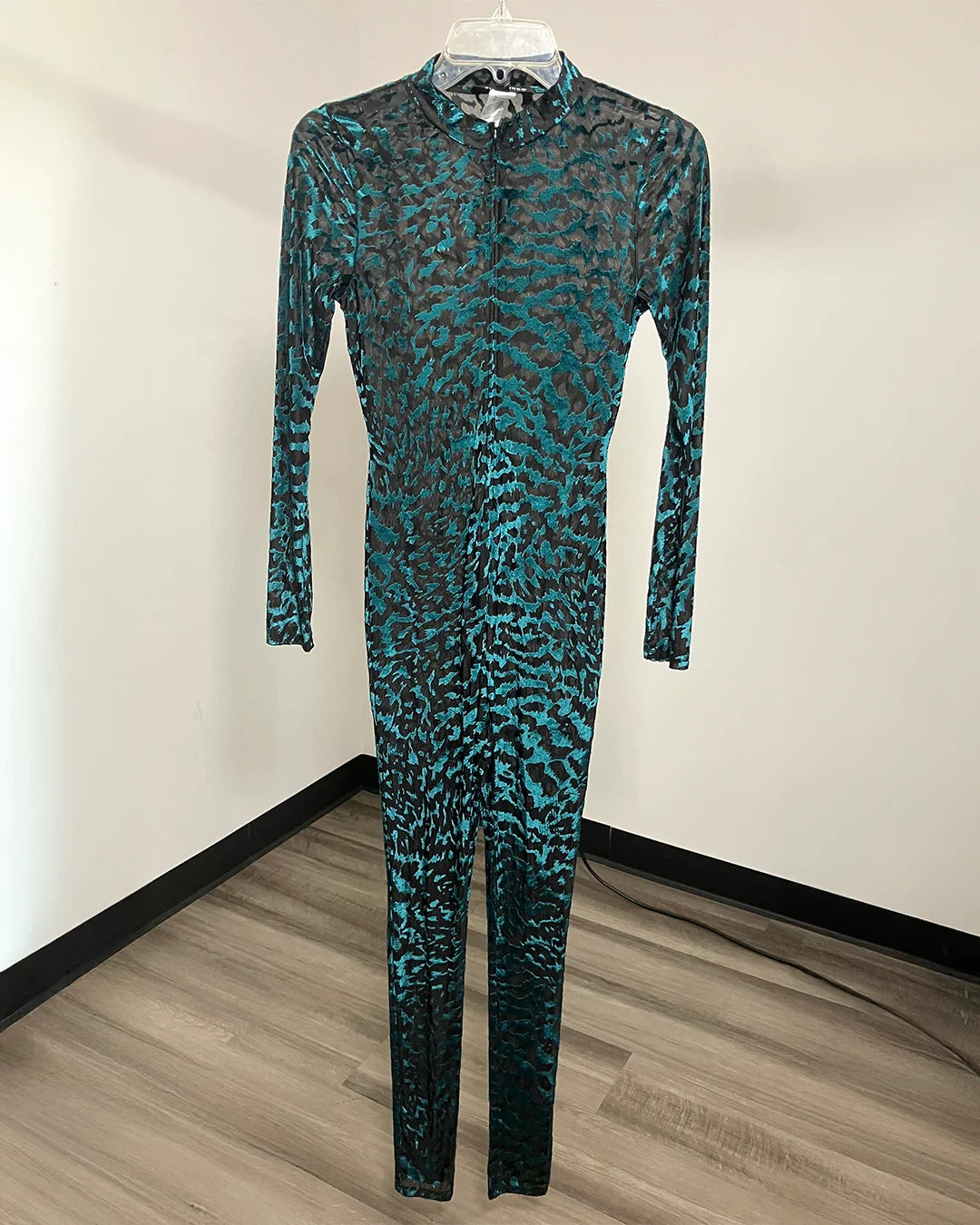 A long-sleeve, full-length one-piece jumpsuit with an abstract teal and black animal-like pattern, hanging on a clear plastic hanger against a plain white wall with a wood-look floor.