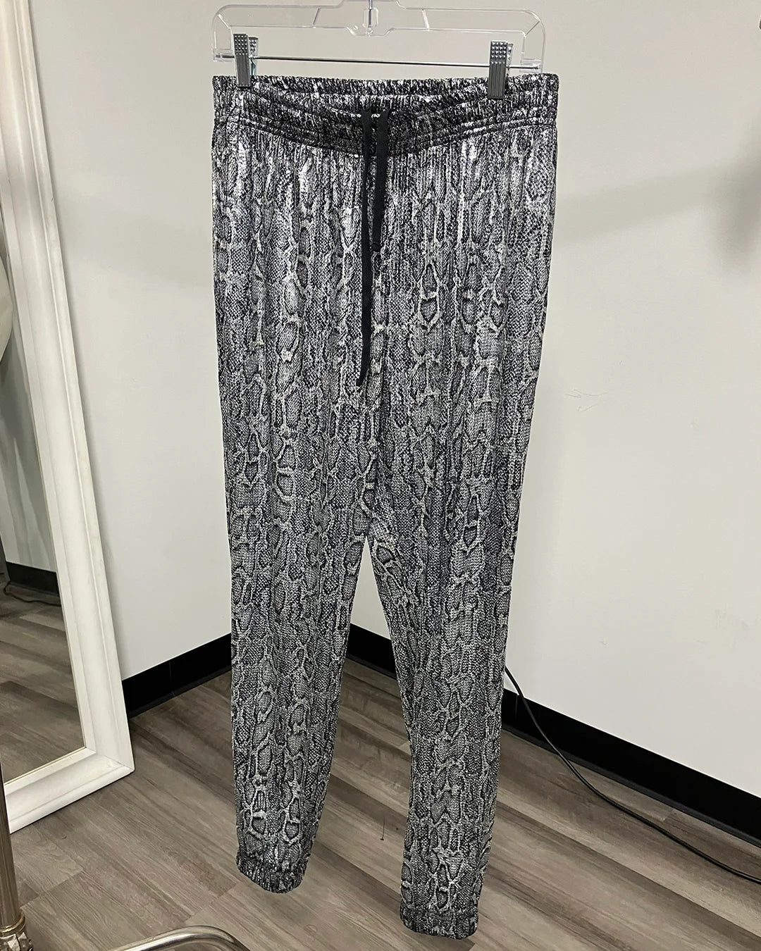 Gray and black metallic snakeskin print joggers hanging on a clear plastic hanger against a white wall.