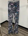 A pair of women's flare pants hanging on a black hanger, featuring a black base with a detailed paisley and floral pattern in white, pink, and light blue, set against an indoor room background.