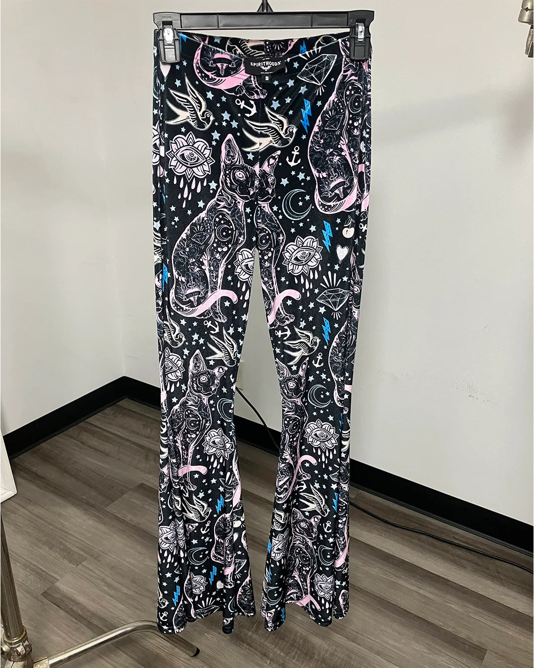 A pair of women's flare pants hanging on a black hanger, featuring a black base with a detailed paisley and floral pattern in white, pink, and light blue, set against an indoor room background.