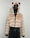 A cropped faux fur jacket with a hood featuring cat ears, displayed on a black mannequin torso. The jacket is a light beige/tan color with black trim and zipper details, paired with black jeans against a plain light gray wall.