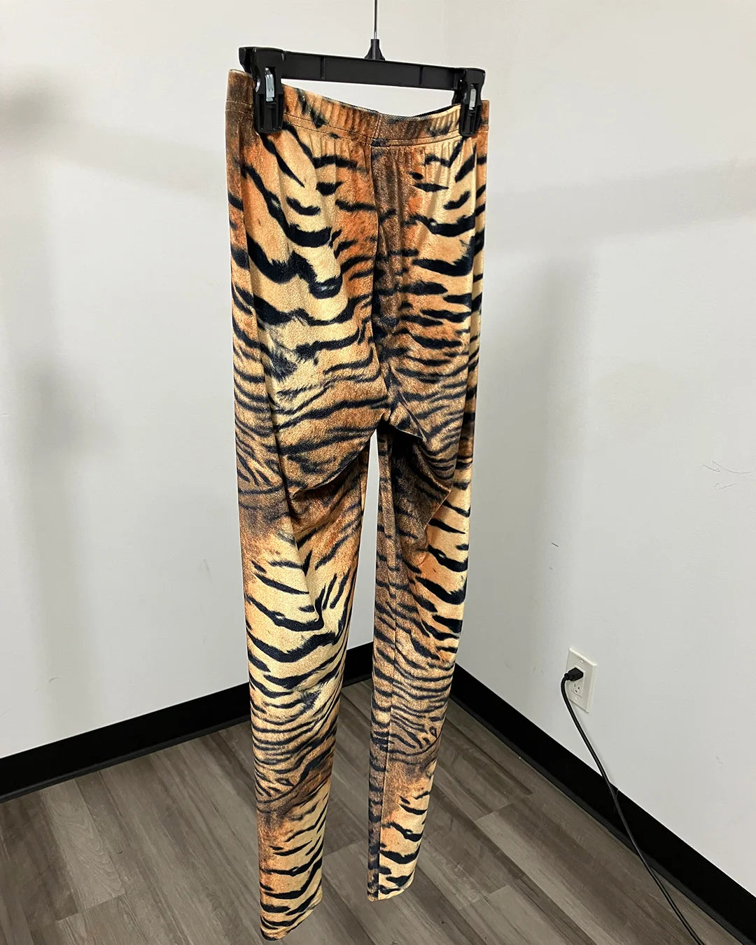Tiger print velvet leggings hanging on a black hanger against a white wall and wood floor.