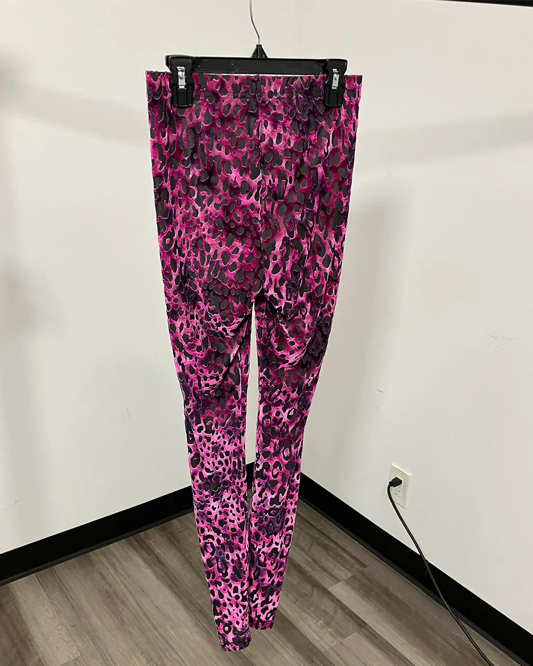 Pink leopard print leggings hanging on a black hanger against a white wall with a wood floor.