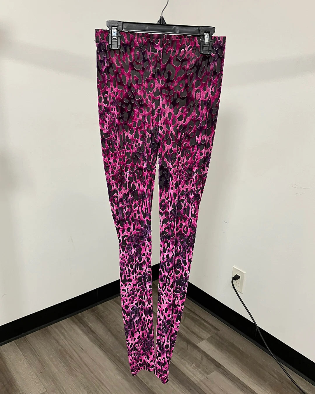 Pink and purple leopard print leggings hanging on a black hanger against a white wall with a wood floor.