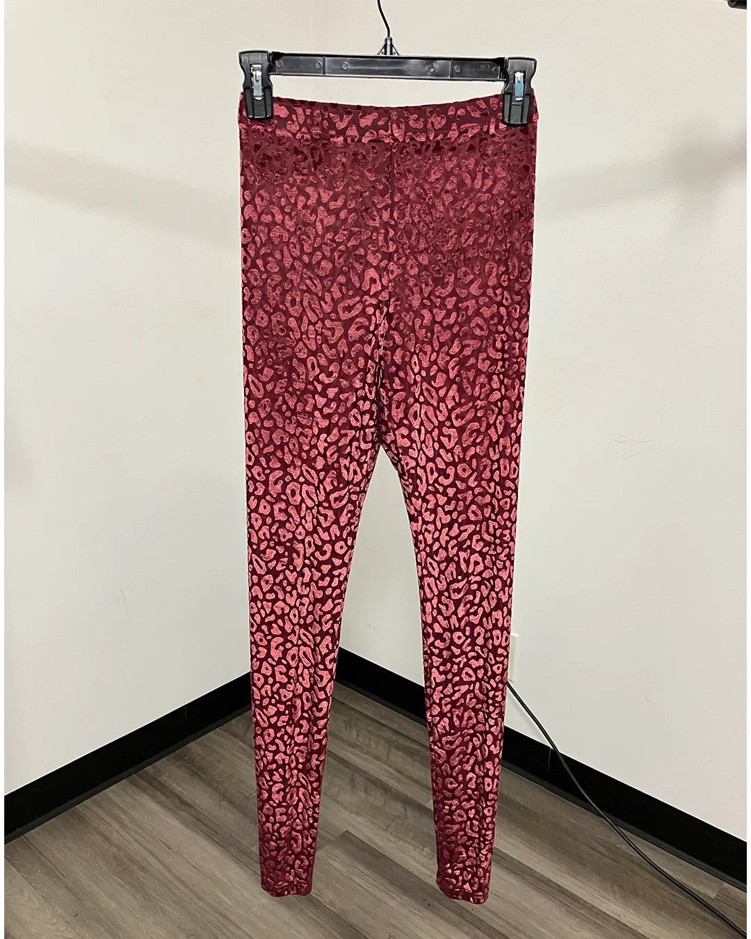 Red leopard print leggings hanging from a black plastic hanger against a plain beige wall and wood-look floor.