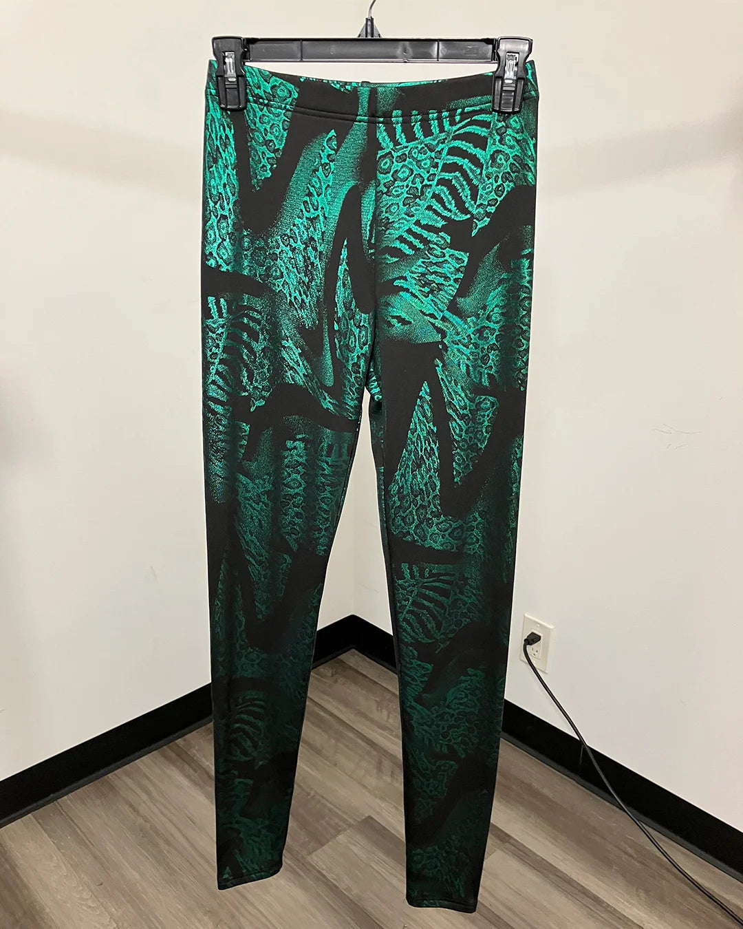 Green patterned animal print leggings hanging on a black hanger against a plain indoor wall with wood flooring visible.