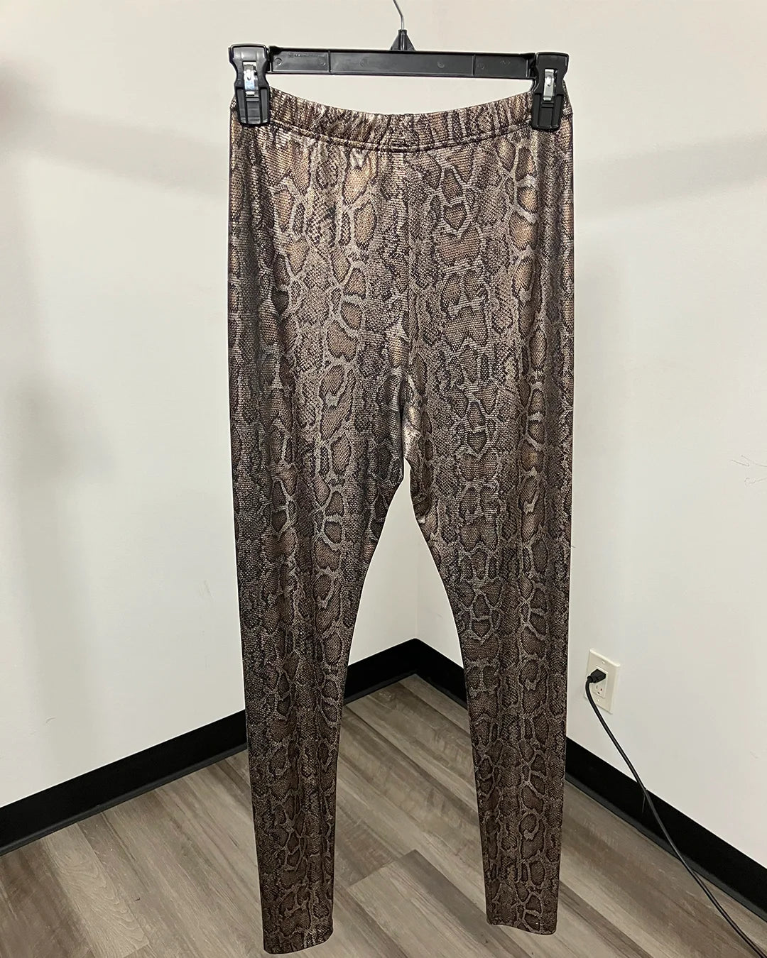 Brown and beige snakeskin print leggings hanging on a black hanger against a plain off-white wall with a wood-look floor.