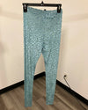 A pair of high-waisted, full-length leggings in a metallic teal blue with a silver leopard print pattern, hanging from a black hanger against a beige wall and wood-look floor.
