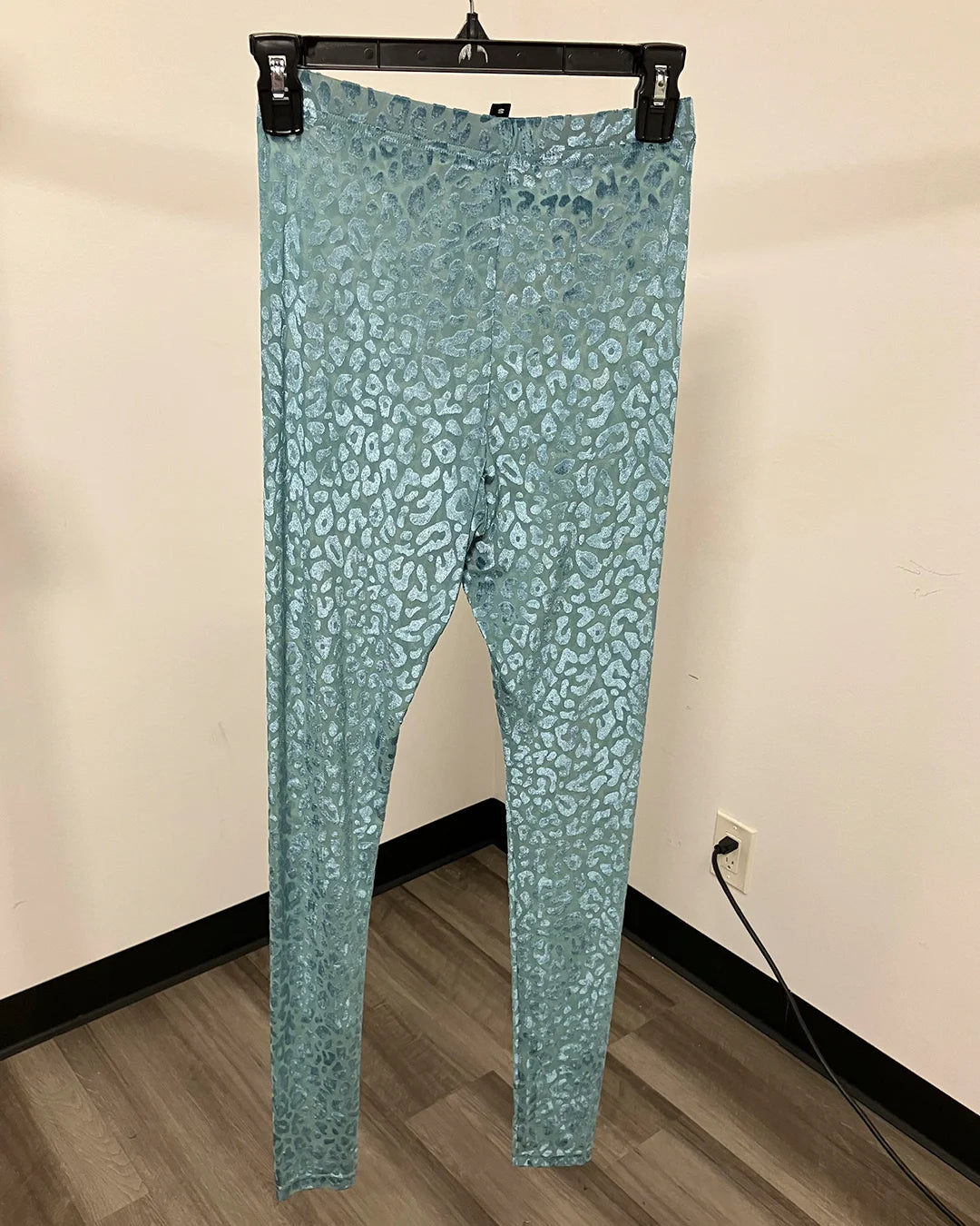 A pair of high-waisted, full-length leggings in a metallic teal blue with a silver leopard print pattern, hanging from a black hanger against a beige wall and wood-look floor.
