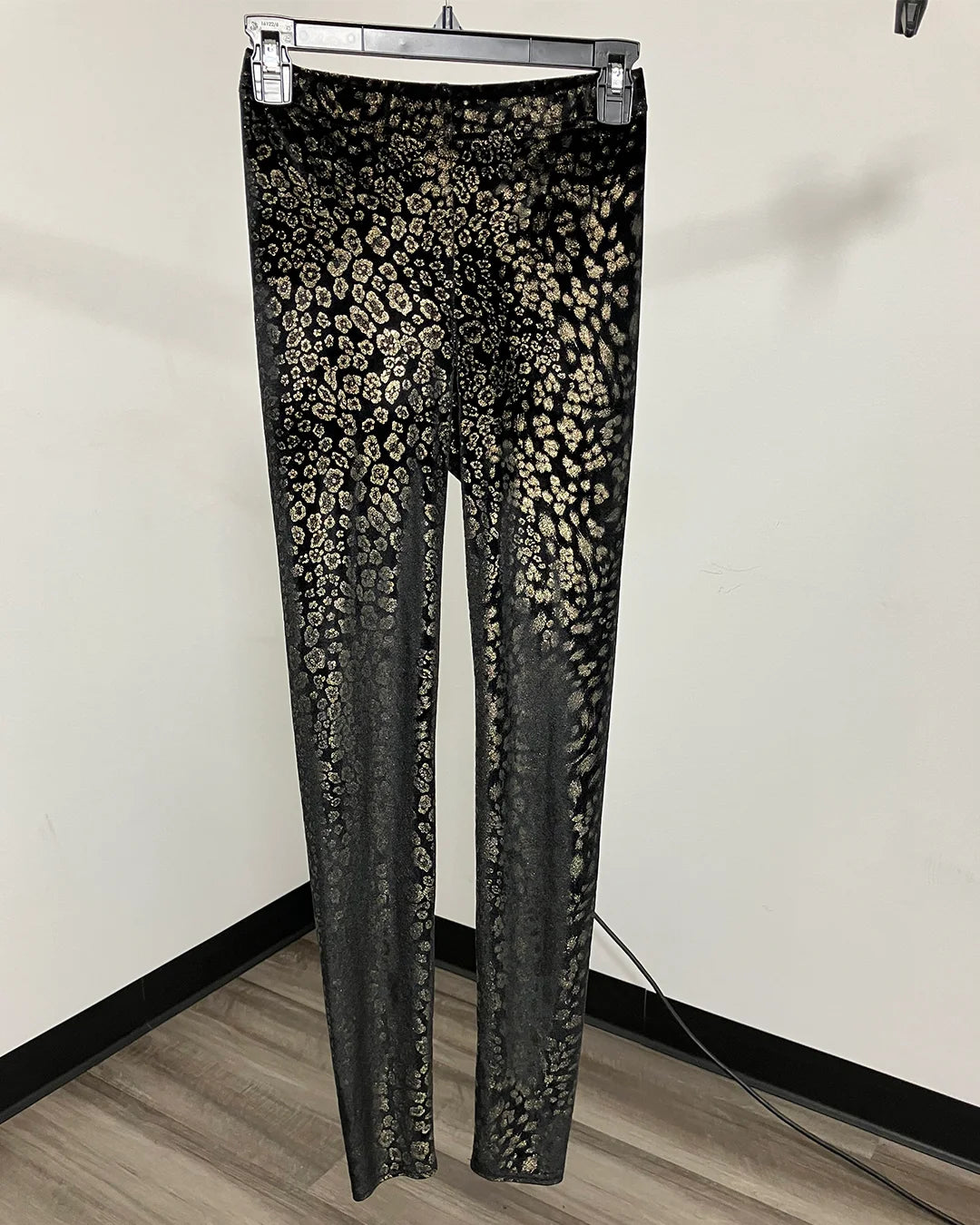 Black and beige leopard print leggings hanging on a metal hanger against an off-white wall and wood floor.
