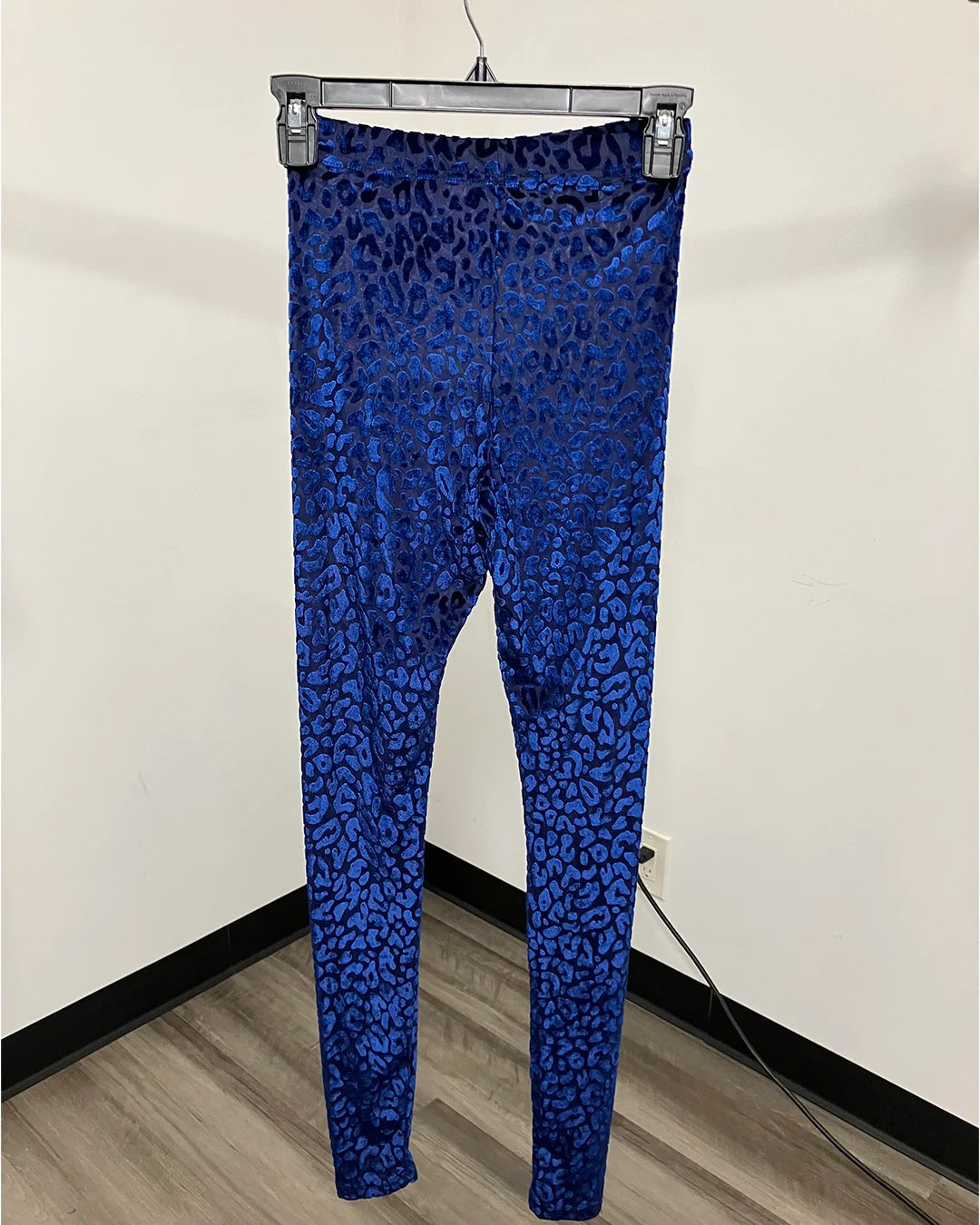 Blue leopard-print leggings hanging on a gray hanger against an off-white wall with a wood-look floor.