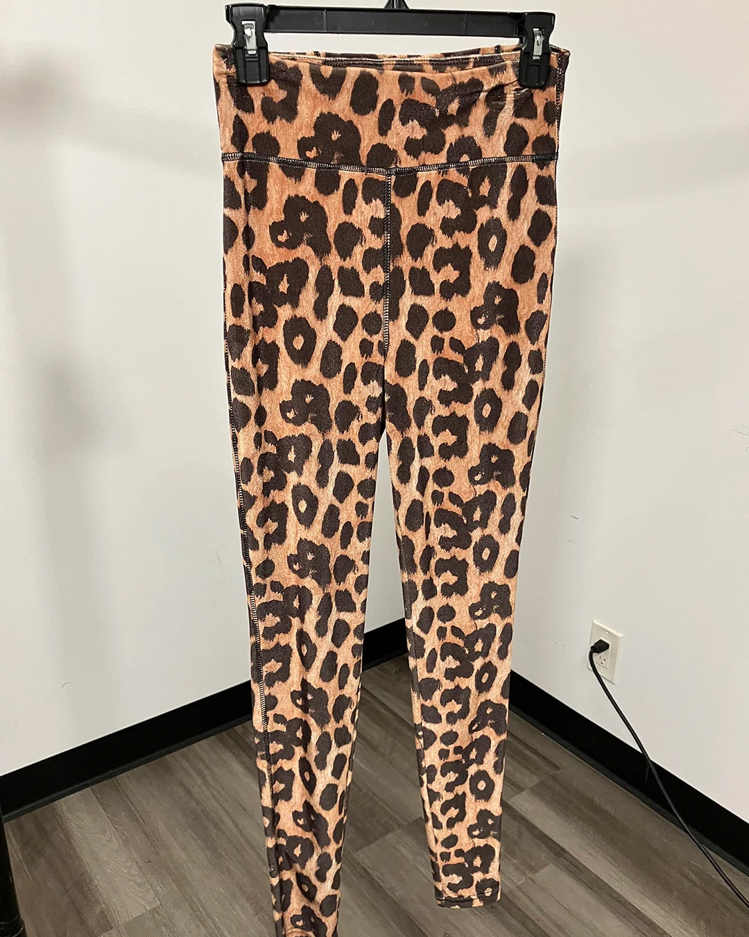 Leopard print leggings hanging on a black hanger against a white wall with a wood floor visible.