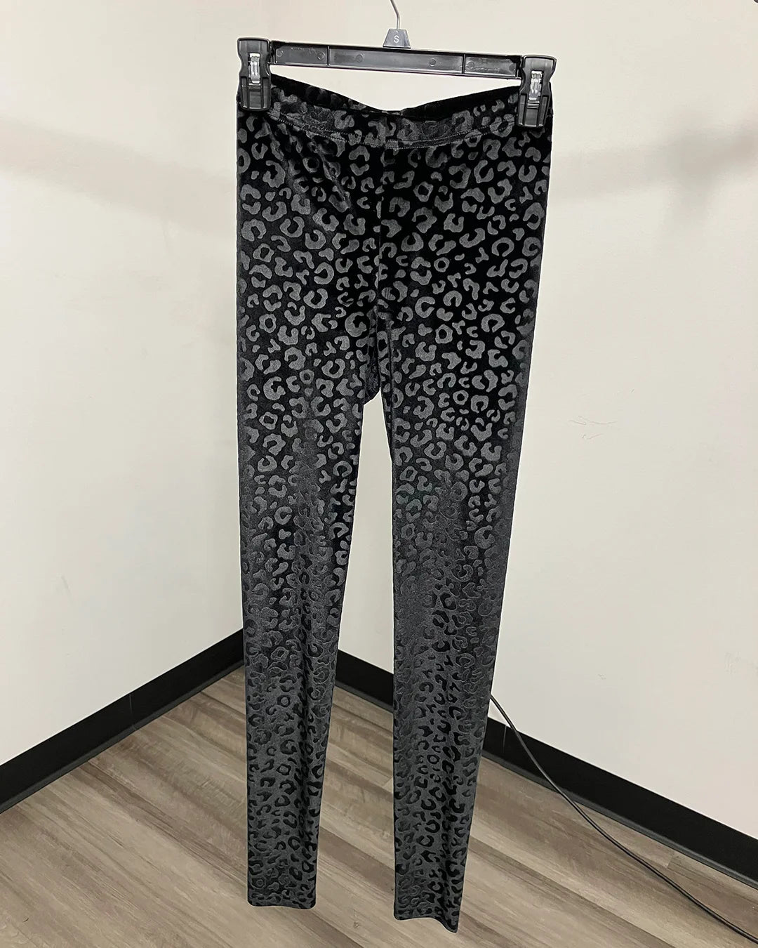 Black leopard print leggings hanging on a black plastic hanger against a plain beige wall and wood floor.