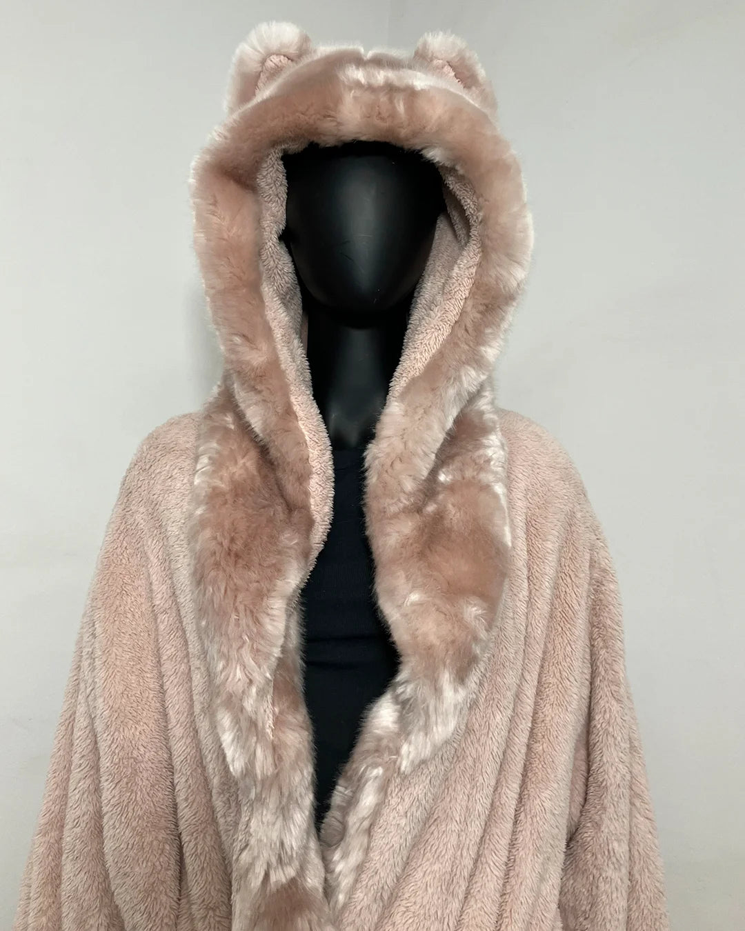 A plush hooded coat in a dusty blush pink faux fur material, featuring ear-like tufts on the hood and a black mannequin torso underneath, set against a plain light gray background.