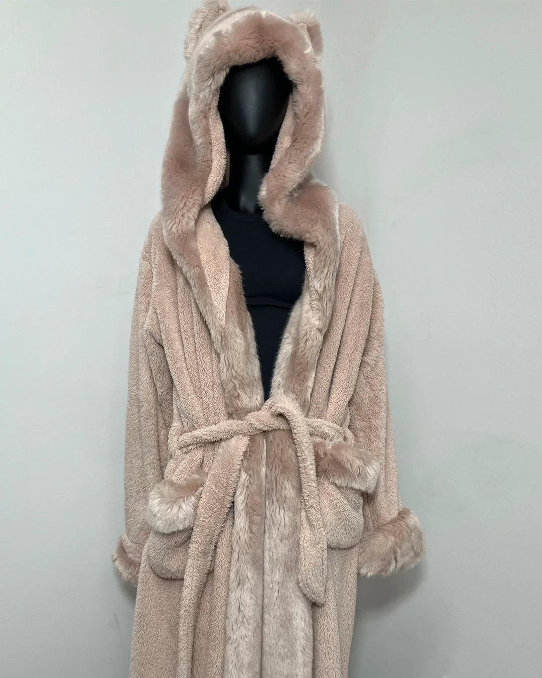 A plush faux-fur hooded robe in a light blush pink color, displayed on a black mannequin torso against a plain light gray wall.