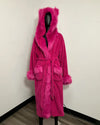 A bright pink plush bathrobe with a matching faux-fur hood, collar, belt, and cuffs, displayed on a black mannequin in an indoor corner setting.