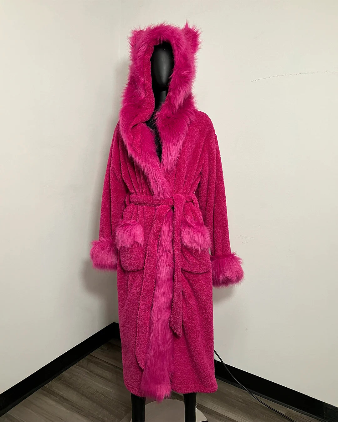 A bright pink plush bathrobe with a matching faux-fur hood, collar, belt, and cuffs, displayed on a black mannequin in an indoor corner setting.