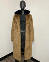 A long tan faux fur coat with a dark navy blue fur-trimmed collar, displayed on a black mannequin torso wearing a black top and dark jeans against a plain white wall.