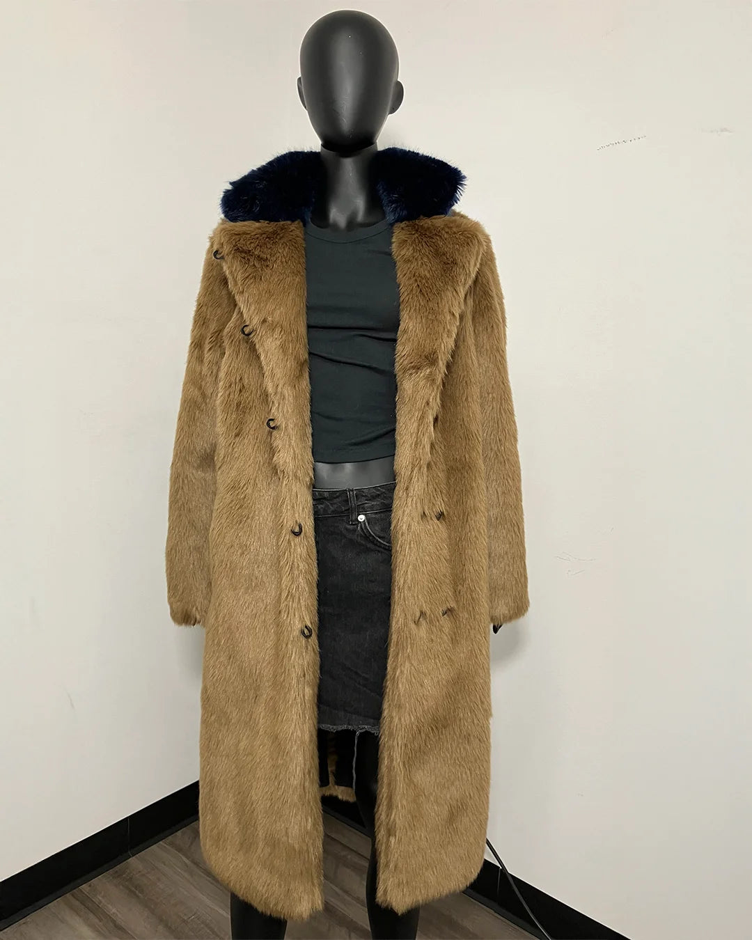 A long tan faux fur coat with a dark navy blue fur-trimmed collar, displayed on a black mannequin torso wearing a black top and dark jeans against a plain white wall.