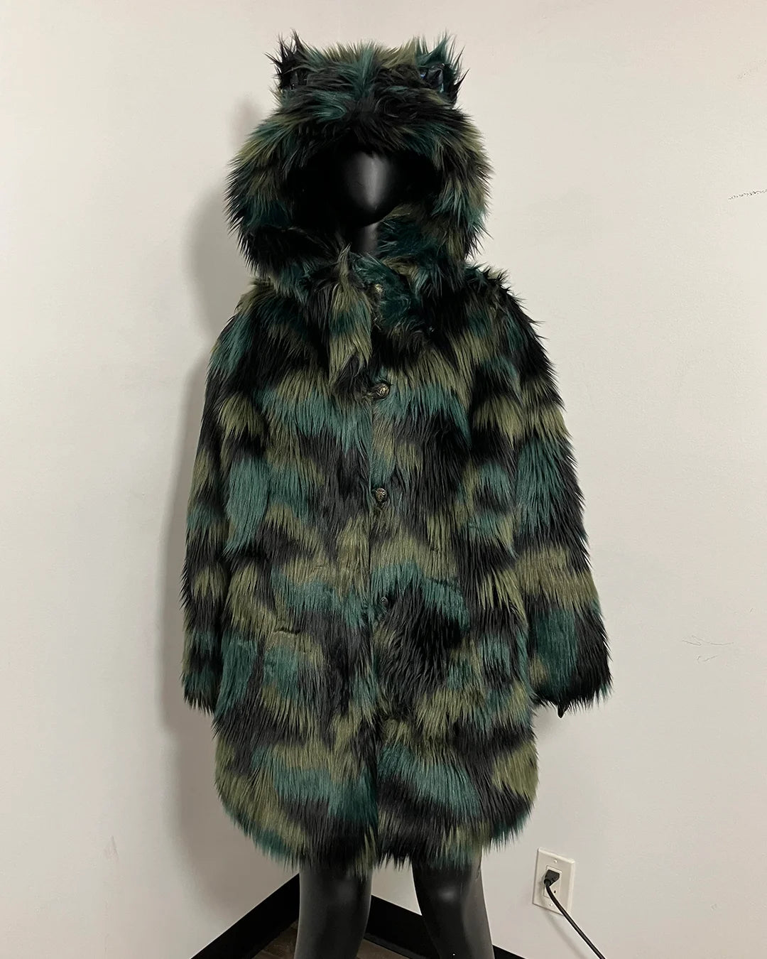 A long faux fur coat with a hood featuring cat-ear tufts, displayed on a black mannequin against a plain off-white wall. The coat has a mottled pattern of black, dark green, and olive-yellow.