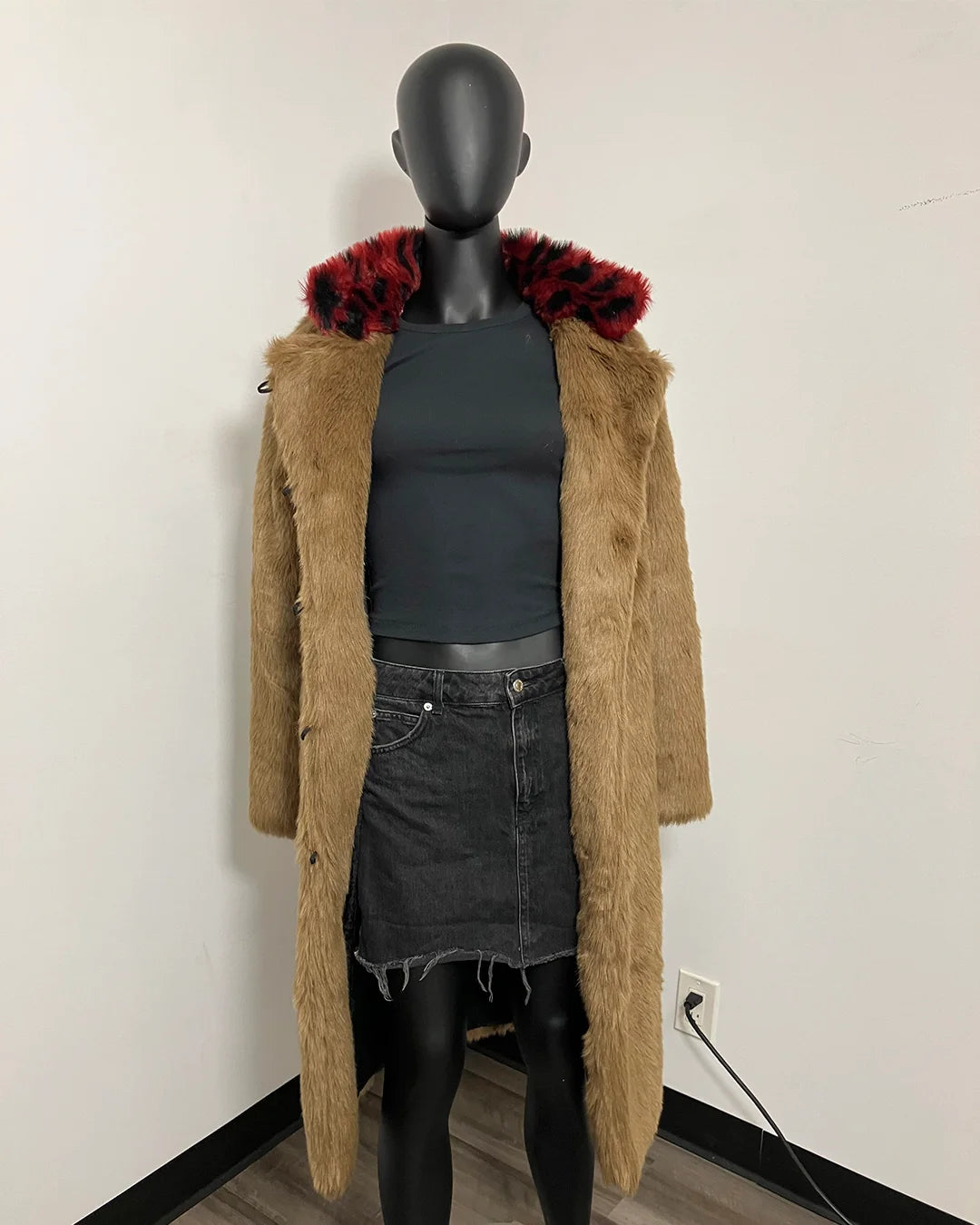 A long tan faux-fur coat with a bold red and black patterned fur collar, displayed on a black mannequin torso wearing a dark gray top and denim skirt against a plain white wall.