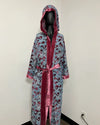 A hooded robe displayed on a black mannequin in an indoor corner. The robe has a light blue base with a dark red and black abstract pattern, trimmed with shiny dark red velvet-like fabric and a matching red tie belt.