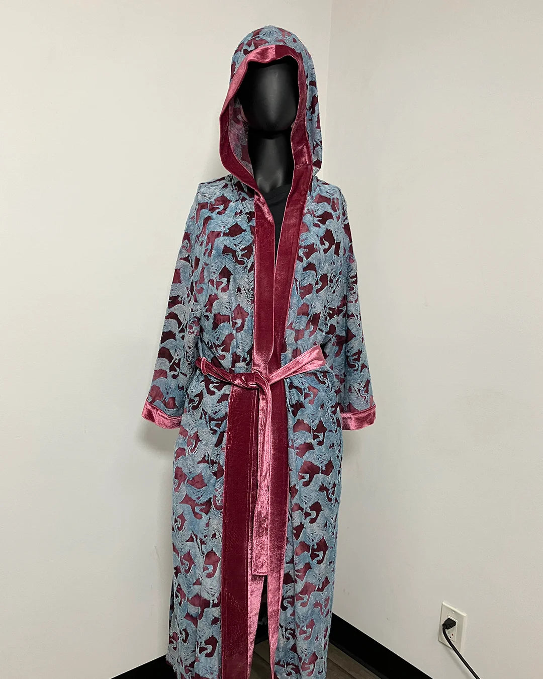 A hooded robe displayed on a black mannequin in an indoor corner. The robe has a light blue base with a dark red and black abstract pattern, trimmed with shiny dark red velvet-like fabric and a matching red tie belt.
