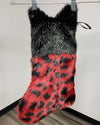 A faux fur Christmas stocking with a black and gray spotted top section and a red and black leopard-spotted lower section, hanging from a metal rod against a plain wall.