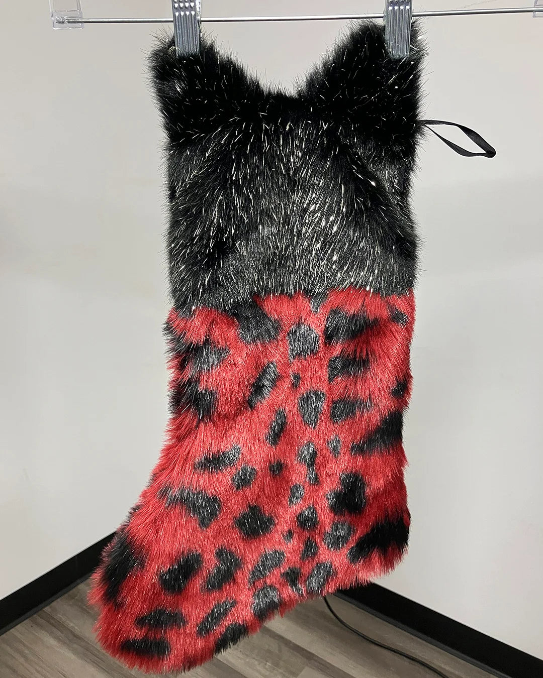 A faux fur Christmas stocking with a black and gray spotted top section and a red and black leopard-spotted lower section, hanging from a metal rod against a plain wall.
