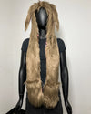 A long, shaggy faux-fur hood in a light tan-brown color with rabbit ears, displayed on a black mannequin torso against a plain off-white wall.
