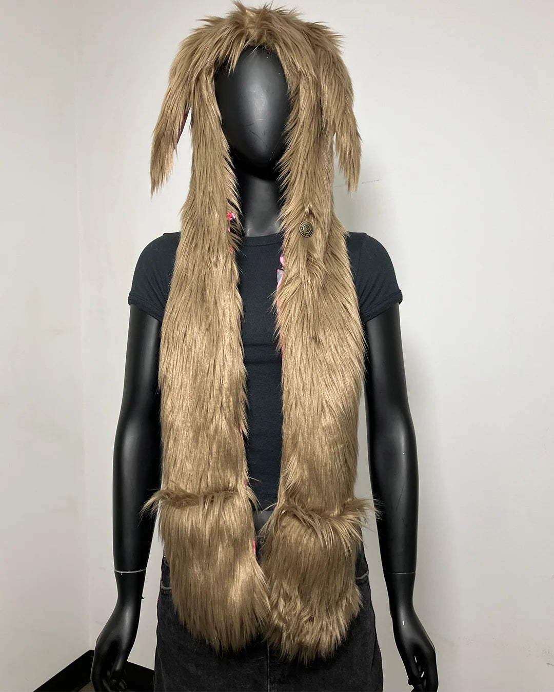 A long, shaggy faux-fur hood in a light tan-brown color with rabbit ears, displayed on a black mannequin torso against a plain off-white wall.