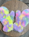 A pair of fuzzy, fingerless gloves in a pastel rainbow tie-dye pattern, displayed on a round wooden surface.