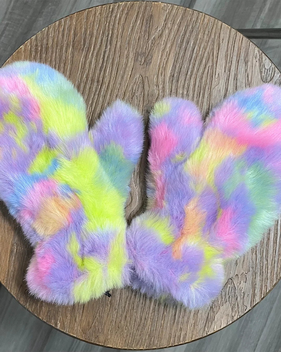 A pair of fuzzy, fingerless gloves in a pastel rainbow tie-dye pattern, displayed on a round wooden surface.