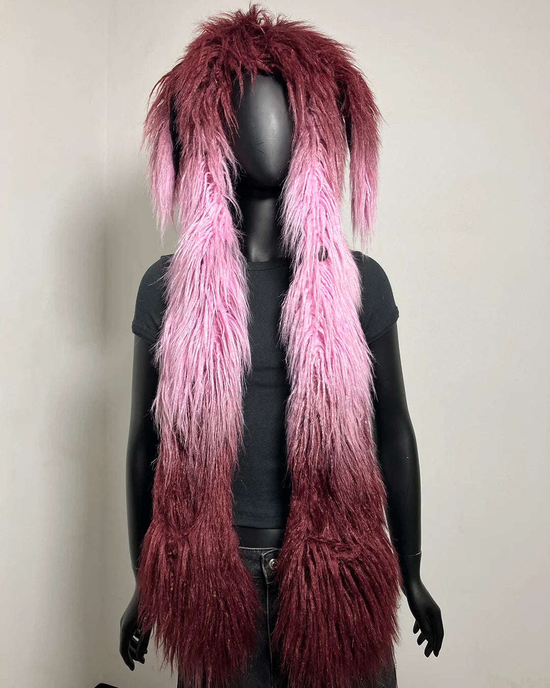 A shaggy, long faux-fur style accessory displayed on a black mannequin torso against a plain light wall. The item has a dark burgundy top section that fades into bright pink at the bottom, resembling ombre hair or fabric.