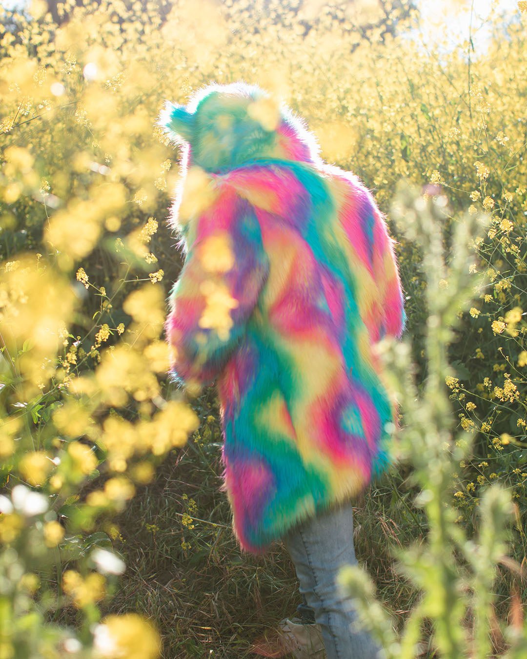 Man in forest of yellow flowers wearing Rainbow Bear Faux Fur Coat with hood and ears up, paired with jeans.