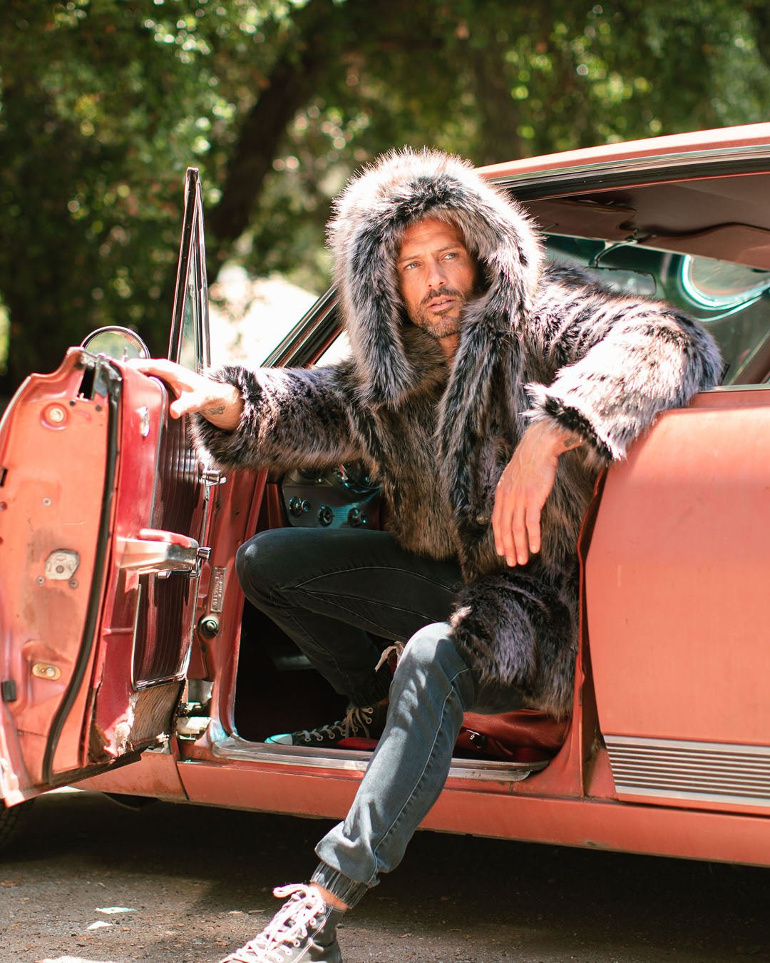 Dude coming out of a coral colored car with one leg out and arm on the side of the window looking away from the camera and wearing the hood up on his grey wolf realistic fur coat.