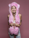 A smiling woman wearing a bright pink faux-fur hooded vest with bear ears and a long matching scarf, posed against a solid dusty pink studio background.