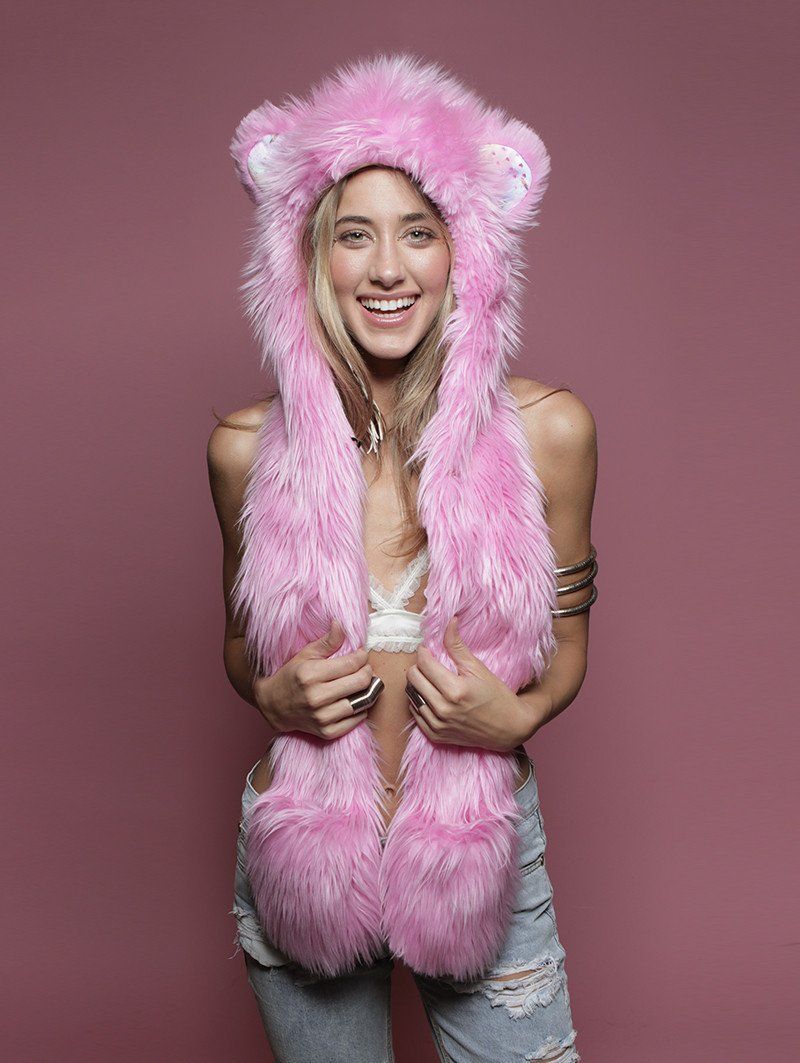 A smiling woman wearing a bright pink faux-fur hooded vest with bear ears and a long matching scarf, posed against a solid dusty pink studio background.