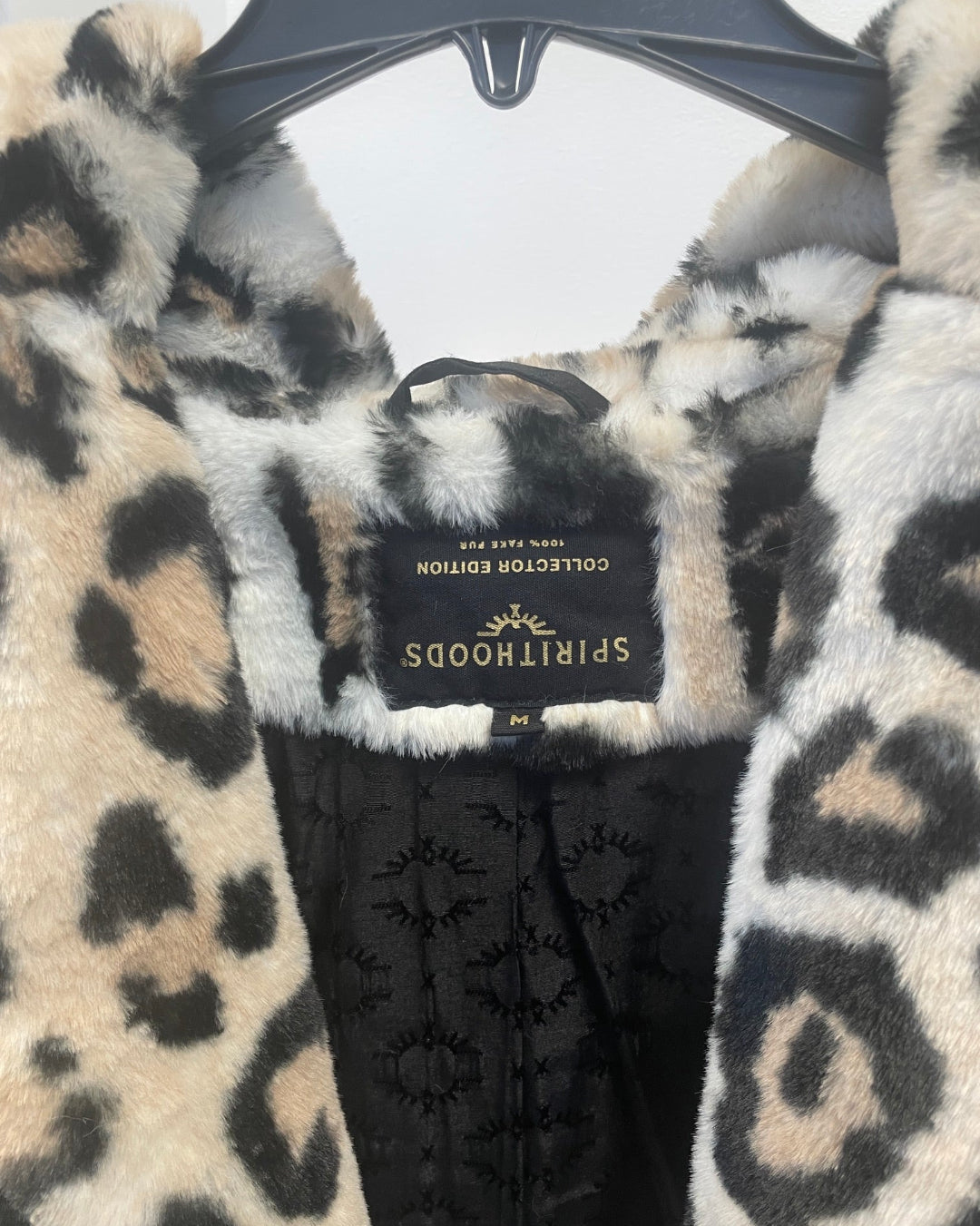 A close-up of a leopard print faux fur jacket by Spirithoods, featuring a black and beige pattern with a black embroidered lining and a black brand label.