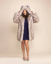 A Woman looking down, wearing a plush luxury grey faux fur coat, holding the sides of an oversized hood, adorned with wolf ears.