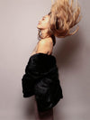 Female brown haired model throwing her hair back and facing one side and wearing the hoodless black faux fur coat draped around her lower arms in just the sleeves midway down her back and waist. 