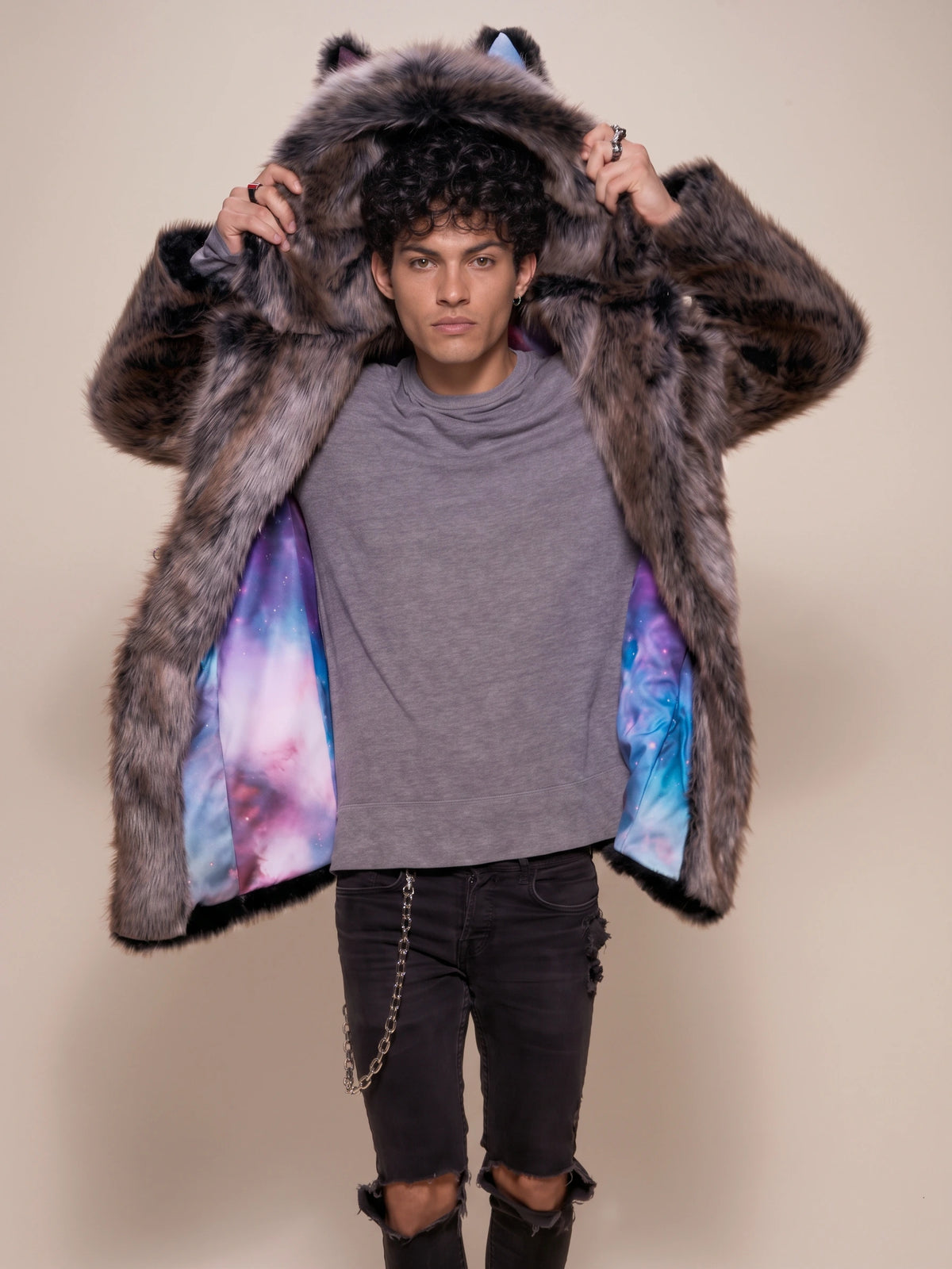 A model wearing a dark brown faux fur hooded coat with colorful galaxy-patterned lining, styled with a gray t-shirt and black ripped jeans against a beige background.